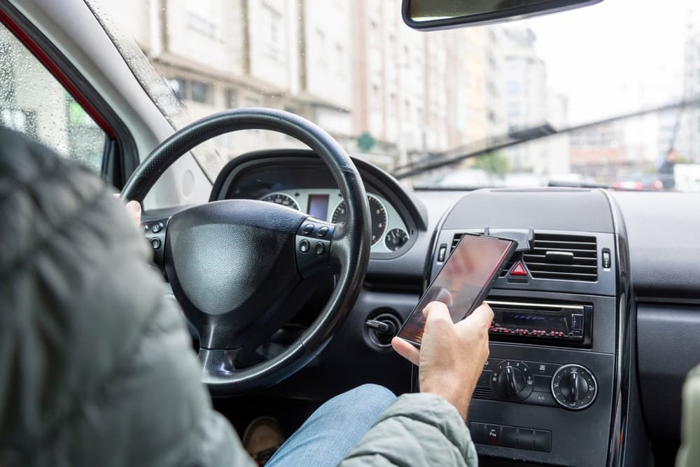 A male driver in a green jacket is dangerously using a smartphone while driving, showing modern distractions and road safety issues