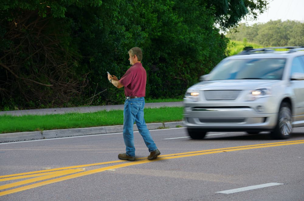 A man who is completely distracted by his cell phone is unaware that he is jaywalking by walking out into heavy traffic on a busy highway road.